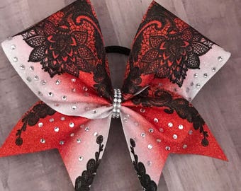May include: A red, white, and black cheerleading bow with a glittery finish. The bow features a lace design and is embellished with rhinestones. The center of the bow is adorned with a silver rhinestone band.
