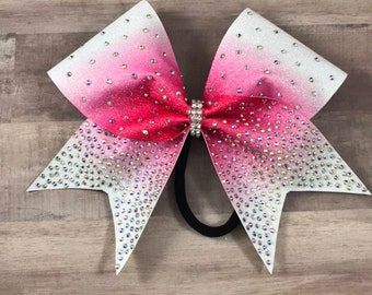May include: A large, glittery cheerleading bow with a gradient of white to pink to red. The bow is adorned with numerous clear rhinestones and has a black elastic band. The center of the bow features a rhinestone embellishment.