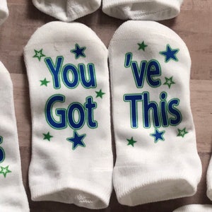 May include: Pair of white ankle socks with blue and green text that reads "You Got This" and "I've Got This". The socks have small green stars printed on them.