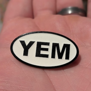 Może przedstawiać: Biała owalna przypinka z czarnym napisem "YEM".
