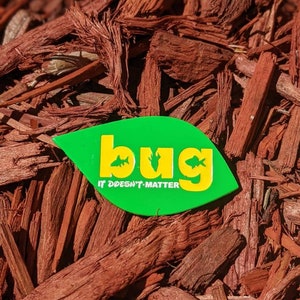 May include: Green leaf-shaped pin with the word "bug" in yellow letters and the text "It doesn't matter" in white letters. The pin is on a bed of red wood chips.
