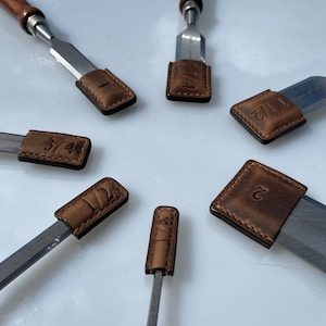 Peut inclure: Un ensemble de huit ciseaux à bois avec des gaines en cuir. Chaque gaine est numérotée de 1 à 8. Les ciseaux sont disposés en cercle sur une surface blanche.