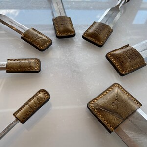 Peut inclure: Six ciseaux à bois avec des gaines en cuir. Les gaines sont brunes et ont un bord cousu. Les ciseaux sont numérotés de 1 à 6. Les ciseaux sont en métal et ont une finition argentée. Les ciseaux sont sur une surface blanche.