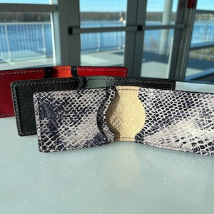 May include: Three leather wallets. The first is red, the second is black, and the third is open, showing a snakeskin pattern. The open wallet has a beige interior. The wallets are on a reflective surface.