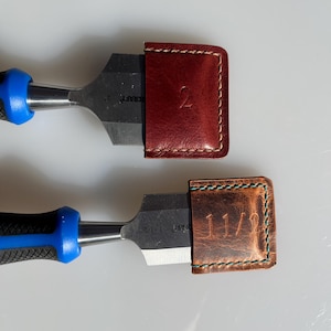 Peut inclure: Deux ciseaux à bois avec des gaines en cuir. Les gaines sont brunes et rouges et portent les chiffres "2" et "1 1/2" en relief.