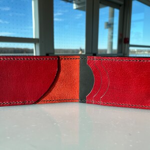 May include: Open leather wallet with a bi-fold design. The exterior is a vibrant red, with black stitching. The interior features orange and black sections for cards and cash. The wallet is open, displaying its compartments.