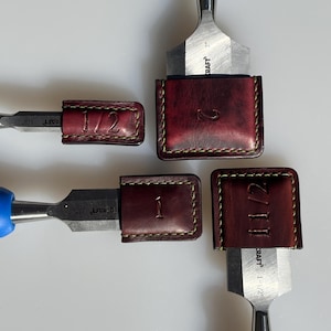 Peut inclure: Quatre étuis en cuir pour ciseaux, chacun avec une taille différente marquée sur le devant : 1/2, 1, 1 1/2 et 2. Les étuis sont de couleur marron avec une teinte rouge et présentent des détails de couture jaunes.