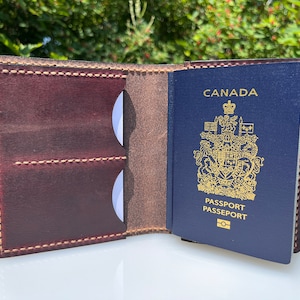 May include: A brown leather passport holder with a stitched design. The holder is open and a Canadian passport is visible inside. The passport is blue with gold lettering and the Canadian coat of arms.