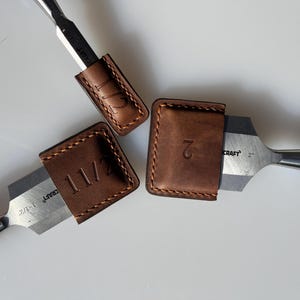 Peut inclure: Trois étuis en cuir pour ciseaux, avec les tailles 1 1/2, 2 et 1/2 pouces gravées dessus. Les étuis sont bruns et ont un bord cousu. Les ciseaux sont argentés et bleus.