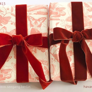 May include: Two rectangular gift packages wrapped in cream-colored paper featuring a repeating coral-pink butterfly and botanical print. Each package is tied with a wide, deep red velvet ribbon finished with a prominent bow. Text reads acajou 415 and havanne 706.