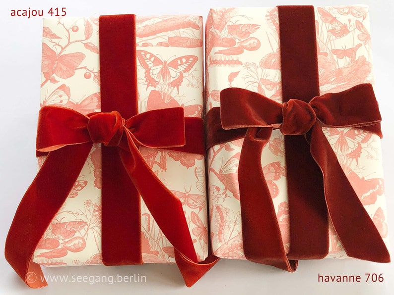 May include: Two rectangular gift packages wrapped in cream-colored paper featuring a repeating coral-pink butterfly and botanical print. Each package is tied with a wide, deep red velvet ribbon finished with a prominent bow. Text reads acajou 415 and havanne 706.