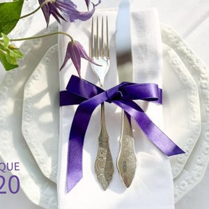 Puede incluir: Una servilleta de lino blanco con una cinta morada atada alrededor de un tenedor y un cuchillo. La servilleta est&aacute; sobre dos platos blancos con un borde decorativo.