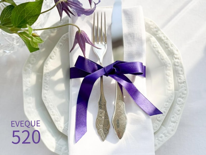 Puede incluir: Una servilleta de lino blanco con una cinta morada atada alrededor de un tenedor y un cuchillo. La servilleta est&aacute; sobre dos platos blancos con un borde decorativo.