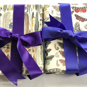 Puede incluir: Dos regalos envueltos con lazos de cinta de sat&eacute;n morado y azul. El papel de regalo presenta un patr&oacute;n de mariposas e insectos sobre un fondo blanco.