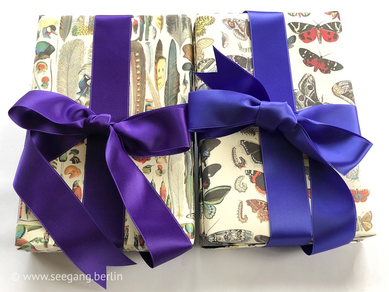 Puede incluir: Dos regalos envueltos con lazos de cinta de sat&eacute;n morado y azul. El papel de regalo presenta un patr&oacute;n de mariposas e insectos sobre un fondo blanco.