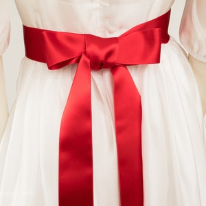 Op de afbeelding: Een witte jurk met een rood satijnen lint dat in een strik is geknoopt op de taille.