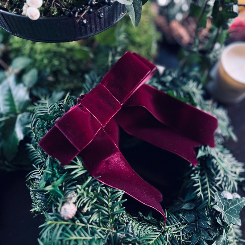 May include: A deep burgundy velvet ribbon tied into a decorative bow, positioned centrally on a circular wreath constructed from dark green evergreen needles and foliage, featuring a smooth, soft texture and a rich, saturated color profile.