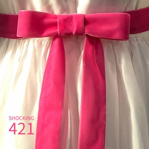 May include: A white dress featuring a vibrant hot pink velvet ribbon tied into a bow at the waist. The fabric of the dress has a textured, layered appearance. The text SHOCKING 421 is printed in pink on the lower left side of the image.