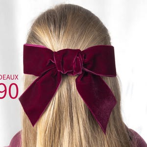 May include: A deep bordeaux velvet hair bow with a knotted center and two long, pointed tails. The text BORDEAUX 390 is printed in red on the left side of the image. The bow is secured in long, straight, light blonde hair.