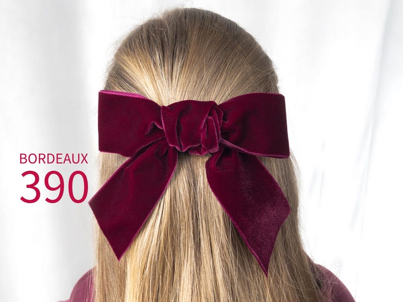 May include: A deep bordeaux velvet hair bow with a knotted center and two long, pointed tails. The text BORDEAUX 390 is printed in red on the left side of the image. The bow is secured in long, straight, light blonde hair.