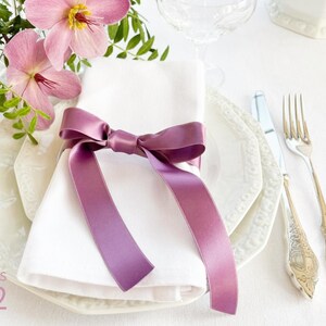 Puede incluir: Una servilleta de lino blanco atada con una cinta de sat&eacute;n morado, colocada sobre un plato blanco con un borde decorativo. El plato est&aacute; sobre un mantel blanco. Un tenedor y un cuchillo de plata est&aacute;n a la derecha del plato. CROCUS 262 est&aacute; impreso en la esquina inferior izquierda de la imagen.