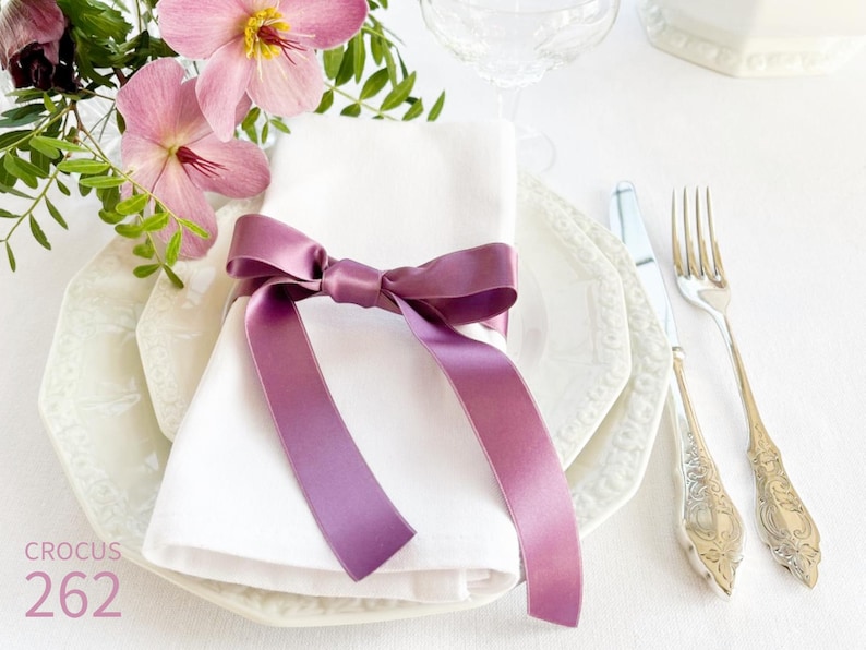 Puede incluir: Una servilleta de lino blanco atada con una cinta de sat&eacute;n morado, colocada sobre un plato blanco con un borde decorativo. El plato est&aacute; sobre un mantel blanco. Un tenedor y un cuchillo de plata est&aacute;n a la derecha del plato. CROCUS 262 est&aacute; impreso en la esquina inferior izquierda de la imagen.