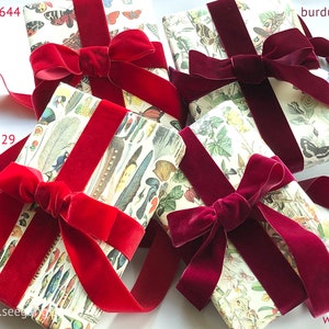 May include: Four rectangular packages wrapped in cream-colored paper featuring botanical and insect illustrations, each tied with a wide velvet ribbon. The ribbons are in shades of scarlet 644, rouge 629, burgundy 434, and wine 424.