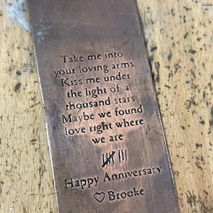 Op de afbeelding: Een koperen bladwijzer met een romantisch gedicht erop gegraveerd. Het gedicht luidt "Take me into your loving arms Kiss me under the light of a thousand stars Maybe we found love right where we are". De bladwijzer heeft ook de tekst "Happy Anniversary Brooke" en een hartje.