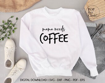 Pode incluir: Camisola branca com texto preto que diz "mama needs COFFEE". A camisola está colocada sobre uma superfície cinzenta com um par de ténis brancos e um par de calças de ganga azuis.