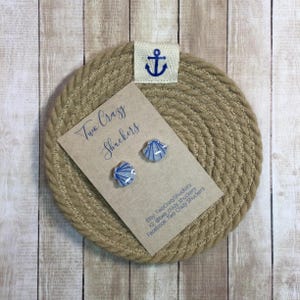 May include: Seashell stud earrings with blue and white patterns, presented on a card reading "Two Crazy Shuckers". The card sits on a woven coaster, with a small tag bearing a blue anchor. The earrings are a charming accessory.