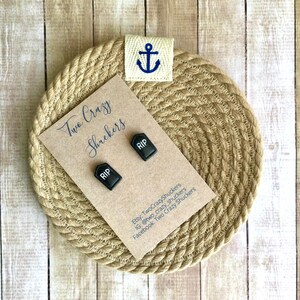 May include: Two black tombstone-shaped earrings with the word "RIP" on them. The earrings are displayed on a brown card with the text "Two Crazy Shuckers" and contact information.