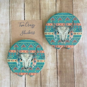 May include: Two turquoise and orange round coasters with a white bull skull and feathers on each. The coasters have a geometric pattern.
