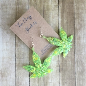 Puede incluir: Un par de pendientes de hoja de marihuana verde y amarilla con ganchos plateados. Los pendientes están hechos de resina y tienen un acabado brillante. Los pendientes están hechos a mano y son una forma divertida y única de mostrar tu amor por el cannabis.