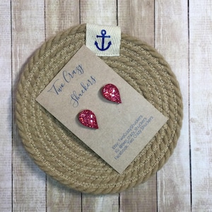 May include: A pair of glittery red teardrop-shaped earrings on a tan card with the text "Two Crazy Shuckers". The card is placed on a round, woven coaster with an anchor emblem.