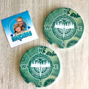 May include: Two round green car coasters featuring a compass design with a mountain and forest silhouette. The text reads NOT ALL WHO WANDER ARE LOST. A square card is included with a photo of two people and the text Two Crazy SHUCKERS.