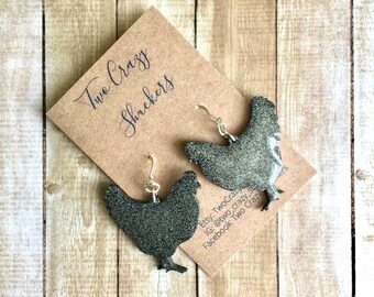 May include: A pair of black glitter chicken earrings with silver hooks. The earrings are on a brown card with the text "Two Crazy Shrekers" written on it.