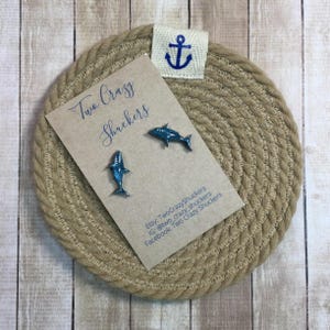 May include: A pair of blue shark earrings on a tan card with the text "Two Crazy Shuckers". The earrings are displayed on a round, woven coaster with a small anchor emblem.