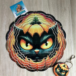 Op de afbeelding: Een ronde metalen windspinner met een zwarte kat met gloeiende gele ogen die uit een uitgesneden oranje pompoen komt, vergezeld van een bijpassende ronde sleutelhanger met hetzelfde ontwerp en een klein vierkant visitekaartje.