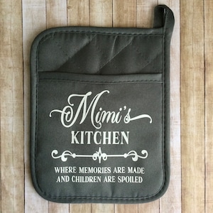 May include: A gray quilted pot holder with a pocket and a hanging loop. The text "Mimi's KITCHEN" is printed in white script, with the phrase "WHERE MEMORIES ARE MADE AND CHILDREN ARE SPOILED" below.