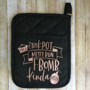 Puede incluir: Manopla de horno negra acolchada con bolsillo y lazo para colgar. La manopla presenta texto en oro rosa que dice "I'm a crock pot messy bun F-BOMB kinda MOM" con un gráfico de una olla, una pancarta y una bomba.