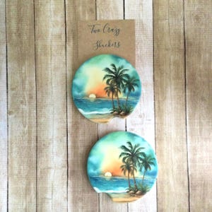 May include: Two round, colorful coasters with a sunset and palm trees design. The coasters are made of ceramic and have a cork backing. The text "Two Crazy Shuckers" is printed on a brown card.