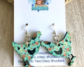 May include: A pair of dangle earrings shaped like chickens on a white display card. The earrings feature a light blue background with a floral pattern and small black and white chicken illustrations. The card includes the text Two Crazy Shuckers.