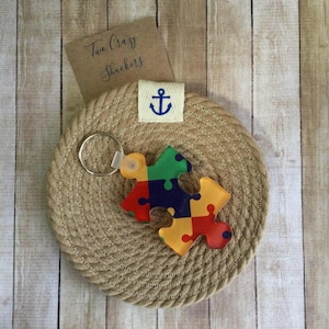 May include: A colorful puzzle piece keychain with red, yellow, green, and blue sections. The keychain is attached to a silver ring and rests on a round, woven coaster. A small tag with an anchor symbol and the text "Two Crazy Shreckers" is also present.