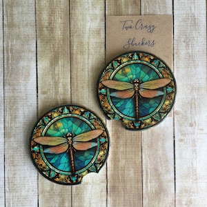 May include: Two round coasters with a stained glass design featuring a dragonfly in shades of teal, gold, and brown. The text "Two Crazy Shuckers" is printed on a brown card.