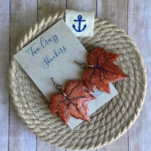Pode incluir: Brincos em forma de folha de cor cobre com detalhes de veias pretas. Os brincos são exibidos em um cartão com o texto "Two Crazy Shuckers" e são colocados em um porta-copos de corda trançada com um emblema de âncora náutica.