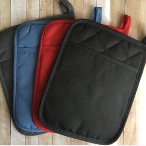 May include: A set of four rectangular quilted pot holders in black, red, blue, and dark gray. Each pot holder features a fabric loop at the top corner for hanging and a stitched diamond pattern on the upper section, providing a textured surface.
