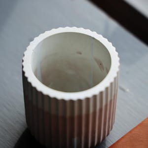 May include: A cylindrical, ribbed ceramic planter with a white scalloped rim. The planter transitions from a light brown to a white interior. A small, brown leather item is visible in the corner. This decorative planter is suitable for small plants.