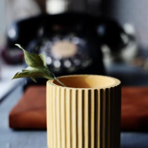 May include: A small, yellow, ribbed ceramic planter with a single green leaf. The cylindrical pot has vertical grooves and a rounded base. A vintage black rotary phone is blurred in the background, along with a wooden cutting board.