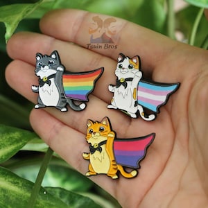 Può includere: Tre spille in smalto con gatti dei cartoni animati che indossano mantelli. I gatti sono grigi, arancioni e calico. I mantelli sono arcobaleno, orgoglio transgender e orgoglio bisessuale.