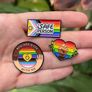 Könnte beinhalten: Drei Emaille-Anstecknadeln in den Farben der Pride-Flagge. Eine rechteckige Anstecknadel trägt die Aufschrift "SAFE PERSON" mit einem kleinen Herz. Eine runde Anstecknadel zeigt zwei Hände, die ein Herz über einer Pride-Flagge halten, mit dem Text "YOU ARE SAFE WITH ME". Eine herzförmige Anstecknadel zeigt eine erhobene Faust.